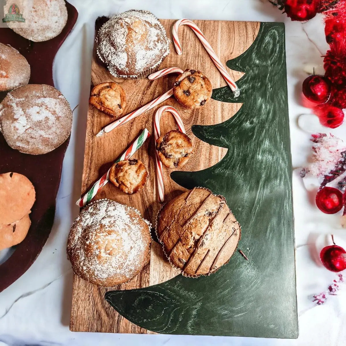 The Christmas Serving Board, hand-crafted and painted with a green Christmas tree, displays muffins, chocolate chip cookies, and candy canes. Additional baked goods and red holiday decorations surround this festive board.