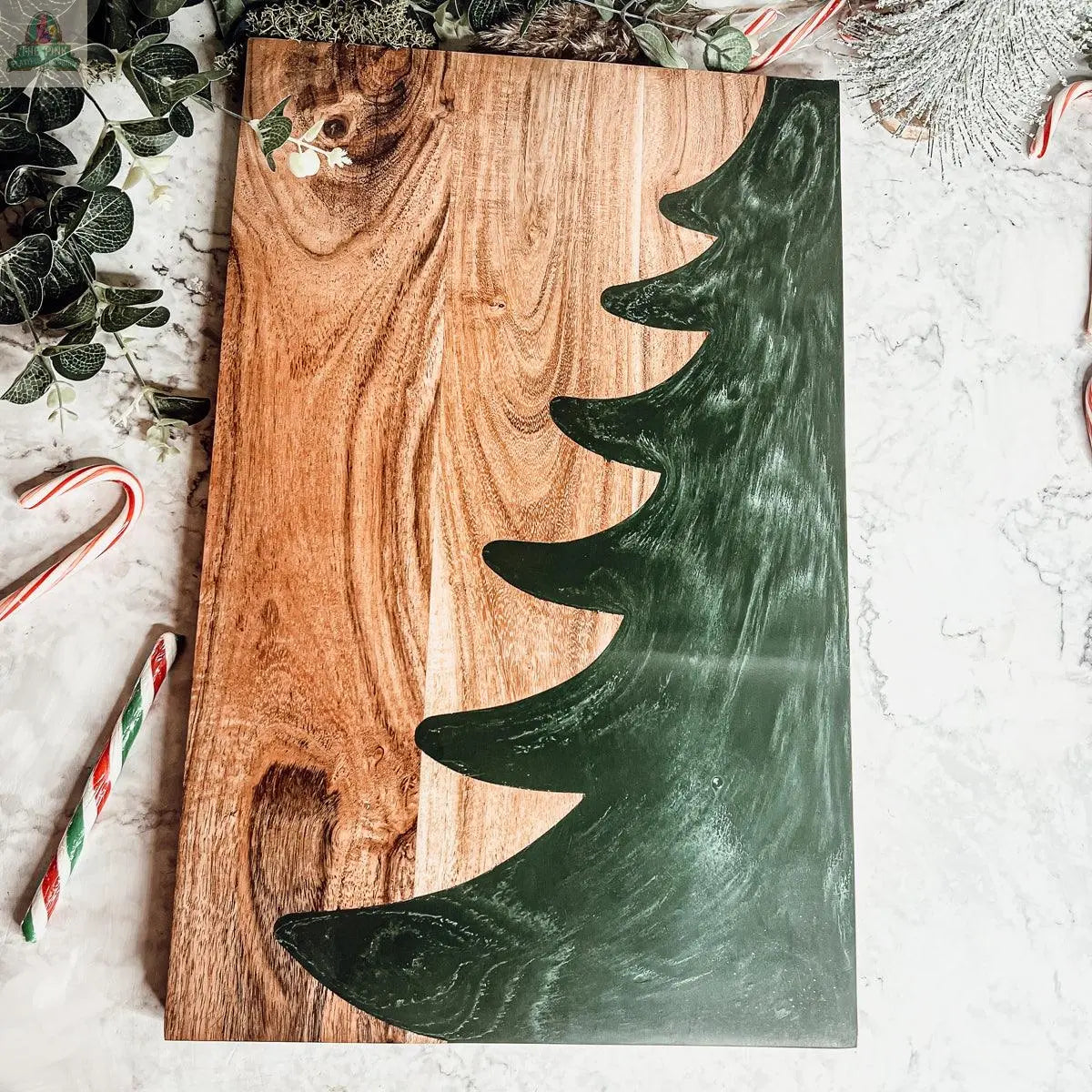 The Christmas Serving Board is a hand-crafted rectangular wooden charcuterie board featuring a green Christmas tree design on one side, surrounded by candy canes and festive greenery on a marble surface.
