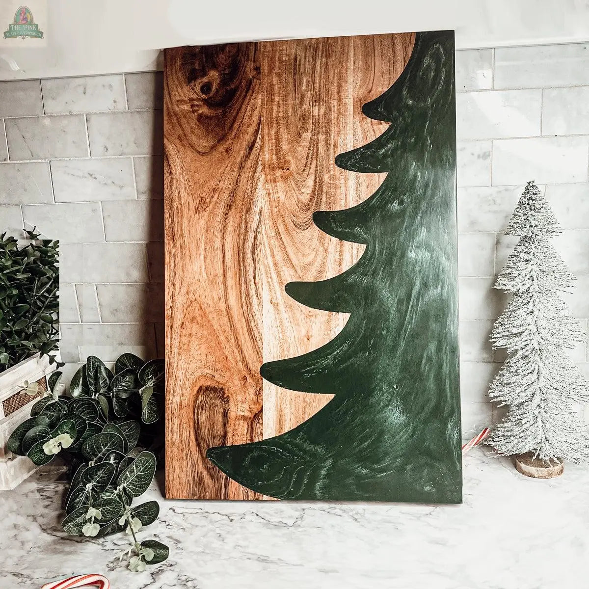 The Christmas Serving Board is a hand-crafted wooden charcuterie board featuring a dark green painted Christmas tree silhouette, displayed on a marble countertop with festive holiday decor and greenery.
