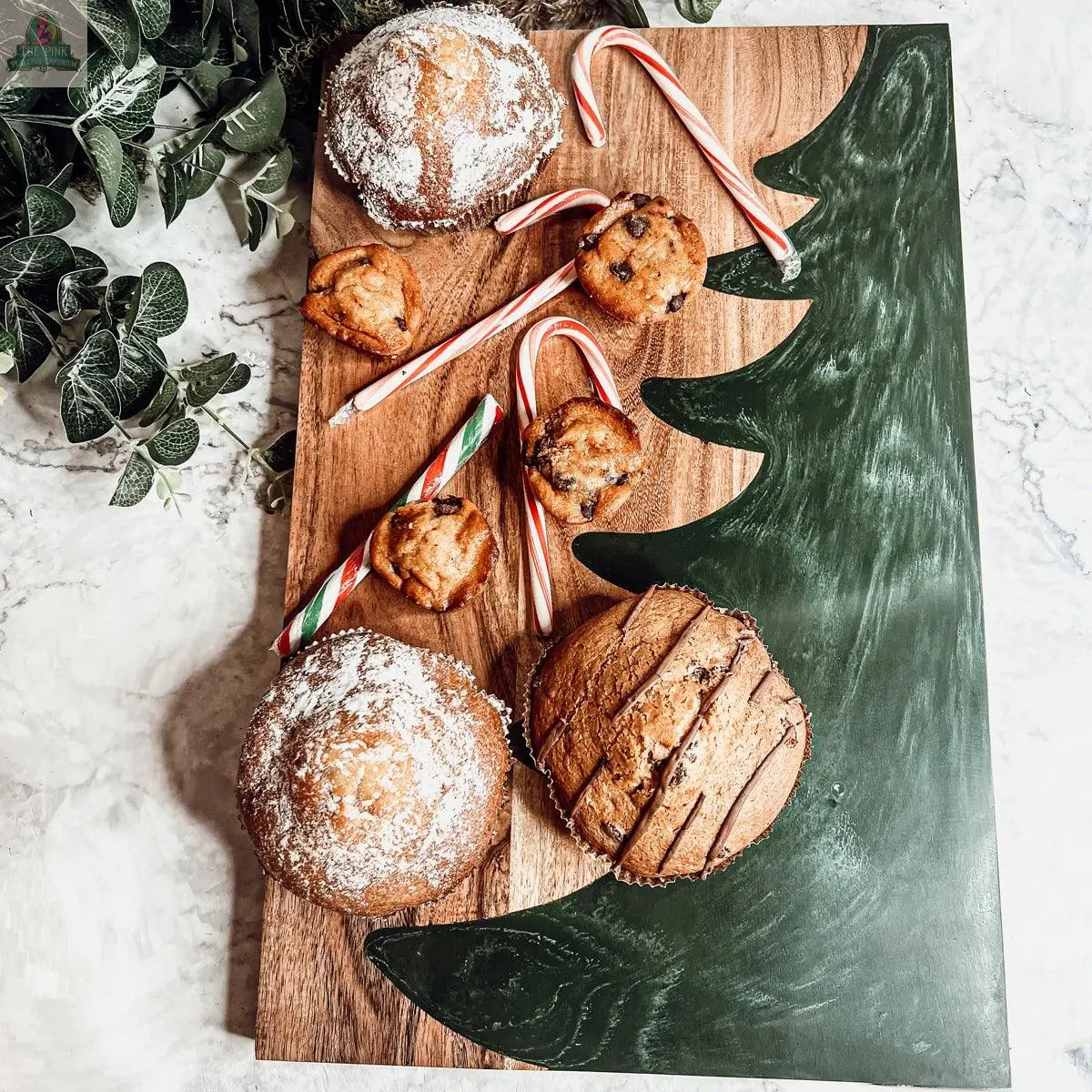 The Christmas Serving Board, featuring a green Christmas tree design, displays muffins dusted with powdered sugar, chocolate chip mini muffins, and three candy canes. Green leaves are seen in the top left corner.