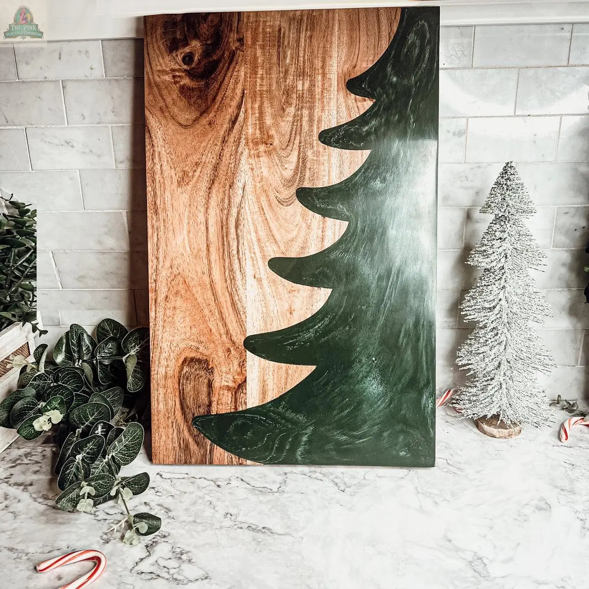 The Christmas Serving Board, a handcrafted wooden piece featuring a green Christmas tree silhouette, is shown on a marble countertop with candy canes, small plants, and a frosted tree, against a white tile backsplash.