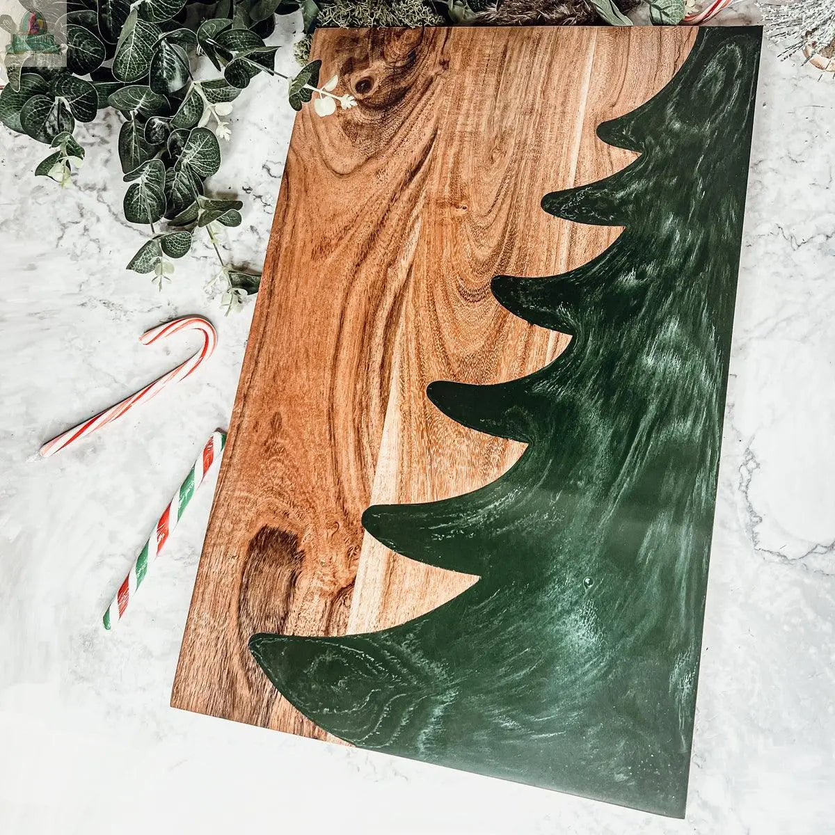 The Christmas Serving Board is a hand-crafted rectangular wooden charcuterie board with a painted green Christmas tree silhouette, displayed on marble with candy canes and green leafy branches.