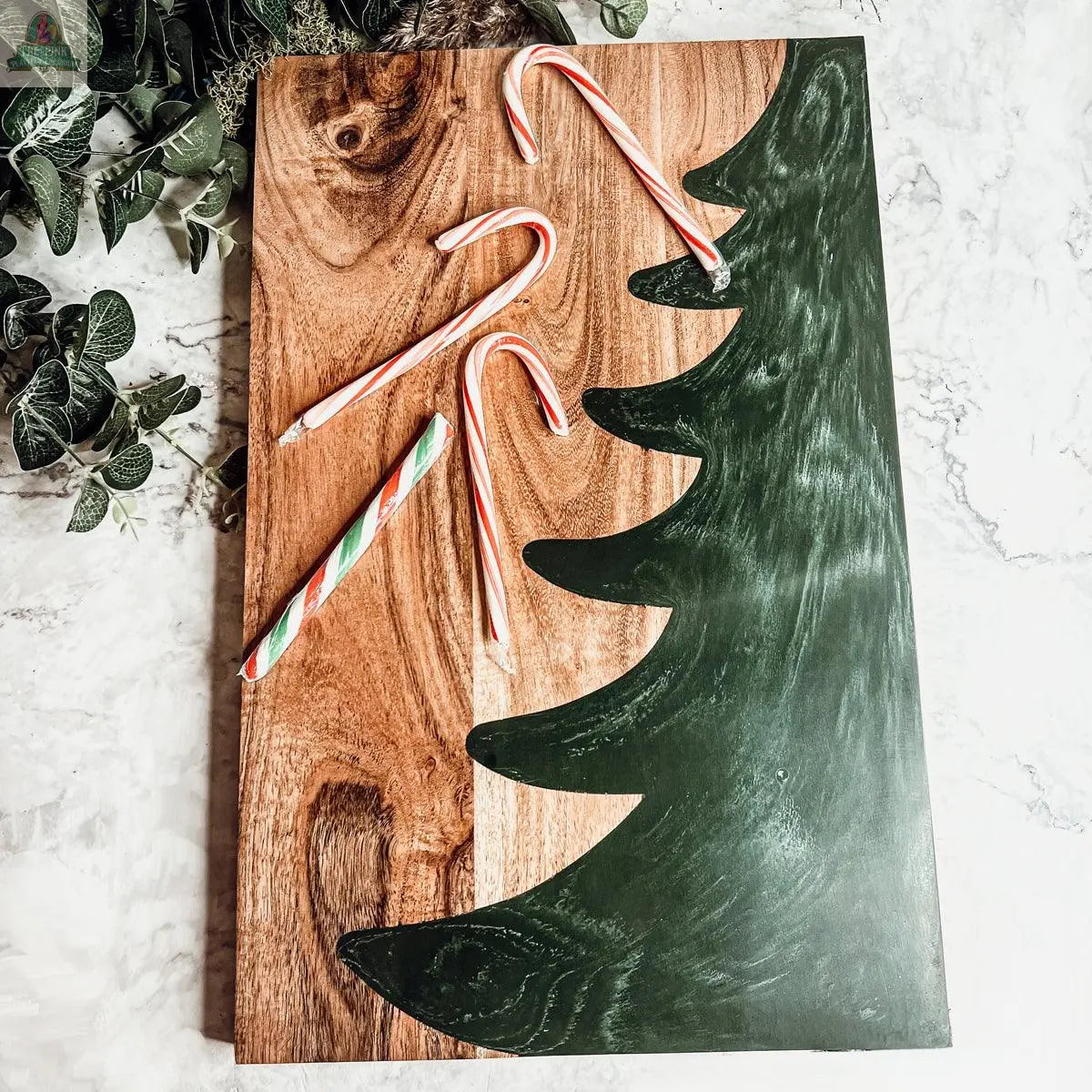The Christmas Serving Board, hand-crafted with a painted green Christmas tree and four candy canes, rests on a light marble surface accented by green leaves in the corner.