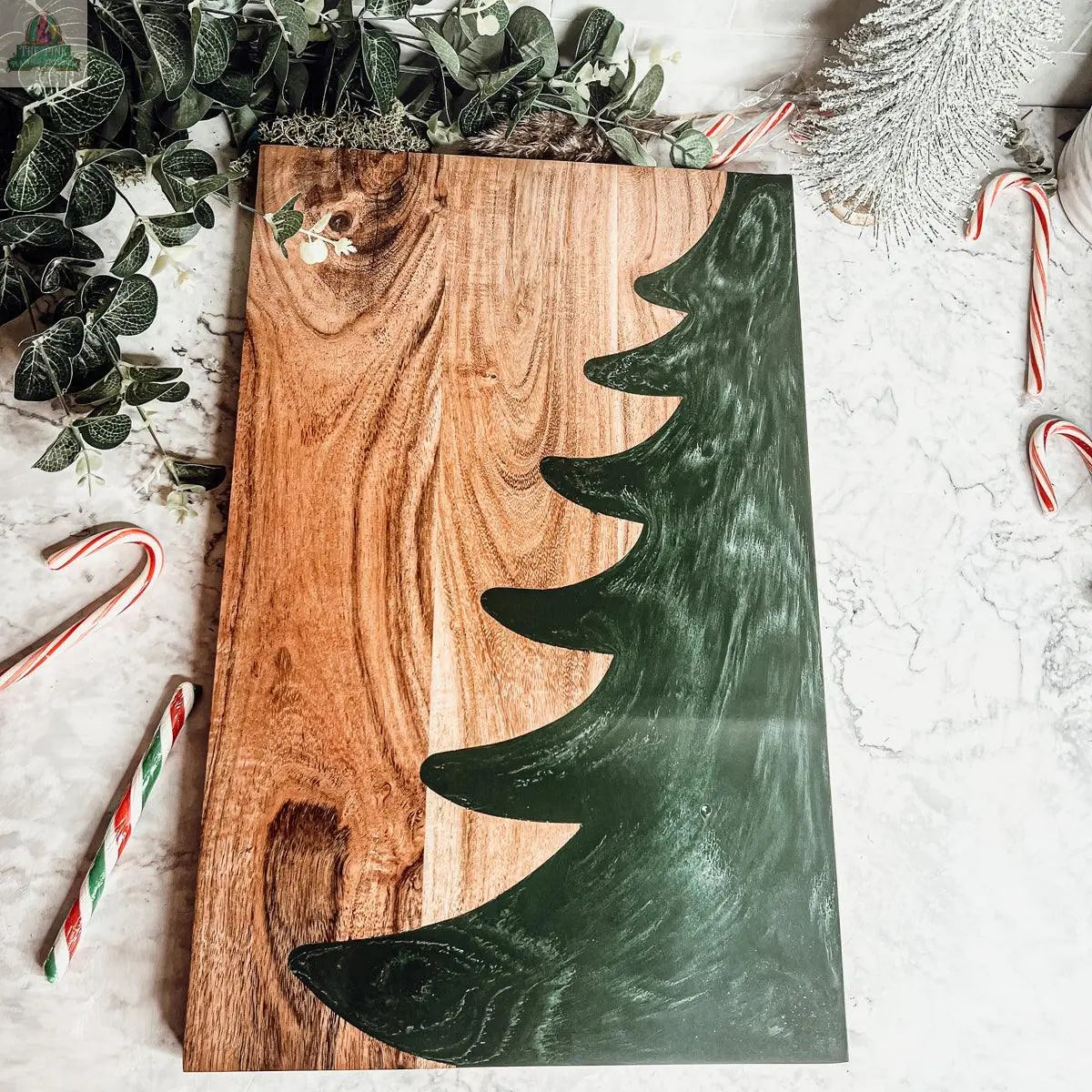 The Christmas Serving Board is a hand-crafted wooden charcuterie board with a green Christmas tree silhouette, surrounded by candy canes, greenery, and a small white tree, displayed on a marble surface.