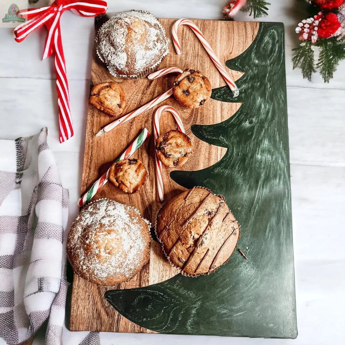 The Christmas Serving Board features a hand-painted green tree, topped with muffins, cookies, and candy canes. Festive accents and a checkered cloth complete this charming holiday centerpiece.