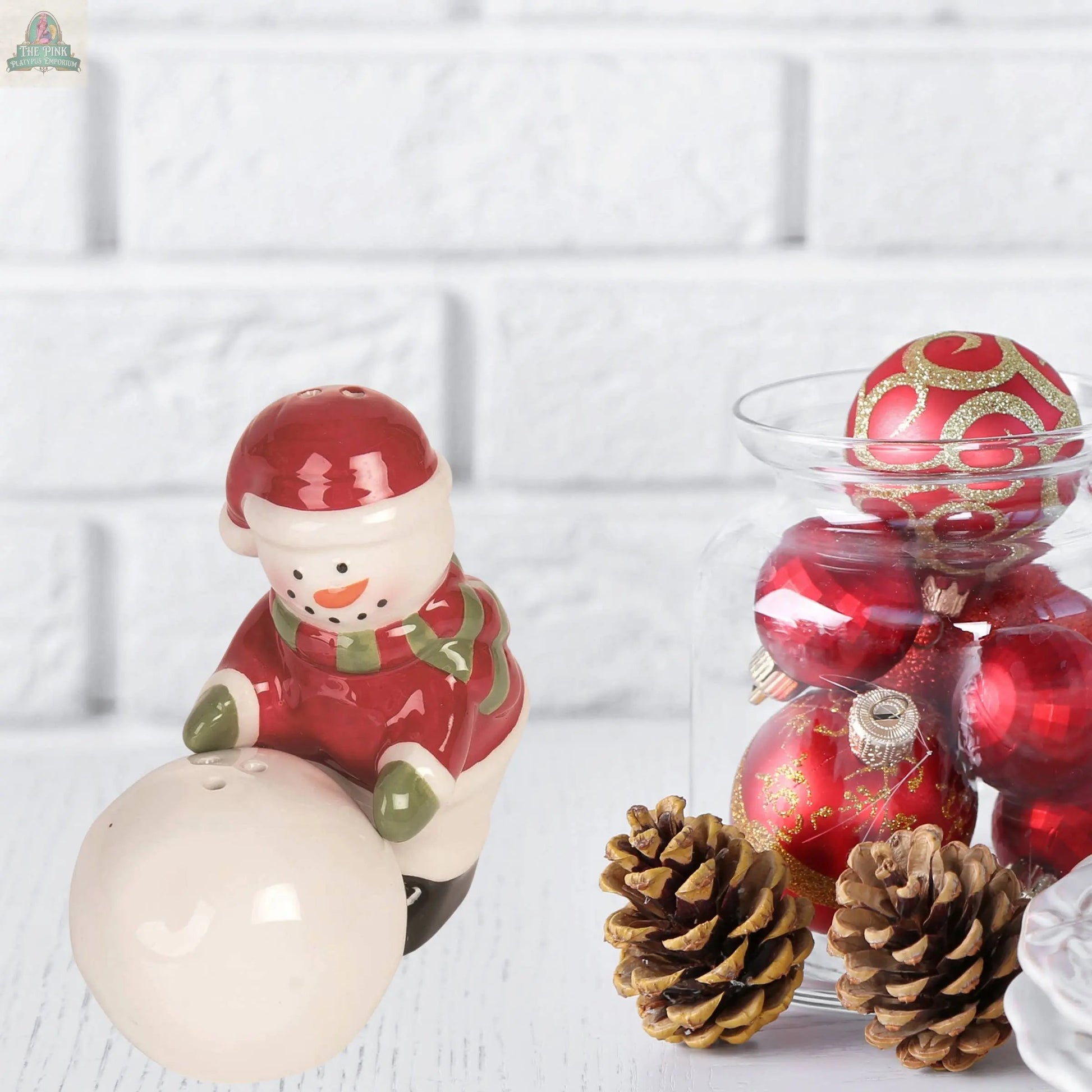 The Christmas Snowman Snowball Salt Pepper Shaker Set (2 pcs, ceramic) in festive red and green sits beside a jar of red ornaments and pinecones against a white brick background—an ideal, charming holiday gift.