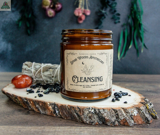 A brown glass jar labeled “Cleansing” sits on a wooden slab, surrounded by sage, a red stone, and black crystals—ideal for energy cleansing. In the background, dried flowers and greenery are visible, softly out of focus.