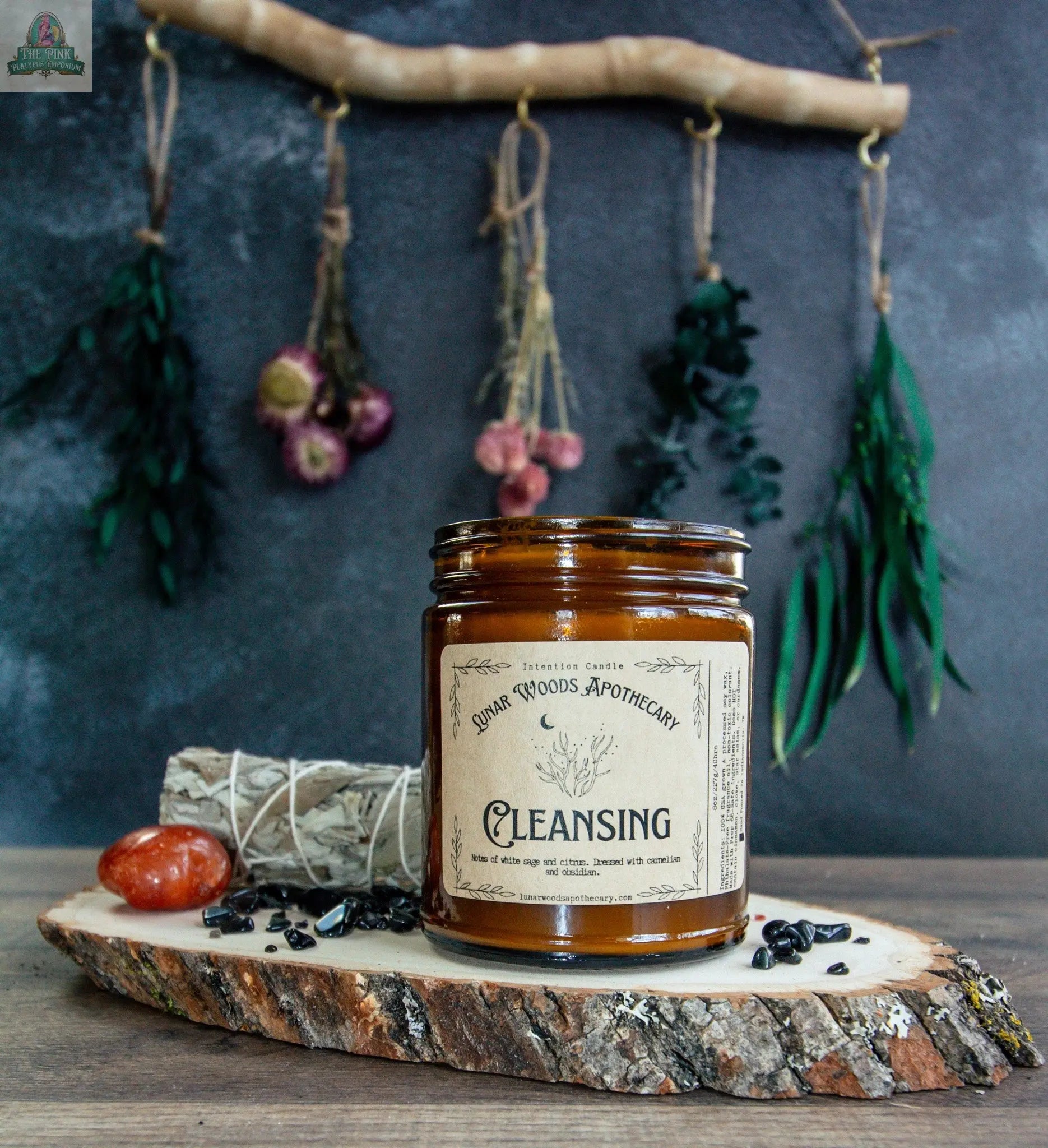 A brown jar of Cleansing rests on a wooden slab, surrounded by a sage bundle, red stone, and black crystals. Dried herbs hang nearby, creating a serene setting ideal for energy cleansing.