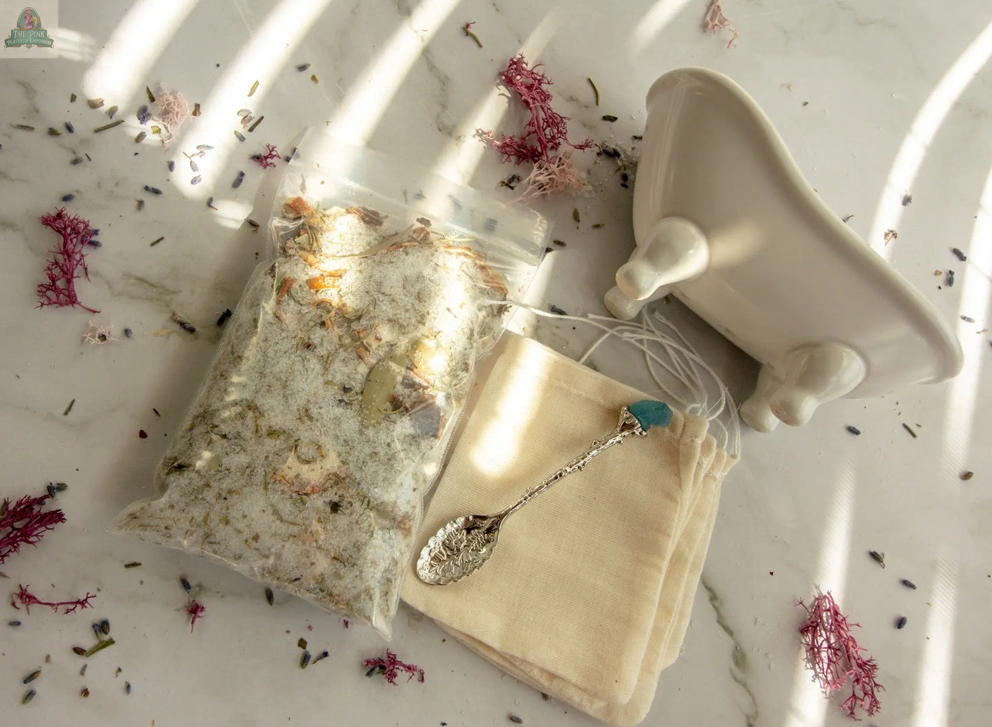 A flat lay of Cleansing bath essentials: a clear bag of herbal bath tea, a floral-patterned silver spoon, a small drawstring pouch, and a mini white ceramic bathtub on marble with scattered flower petals.