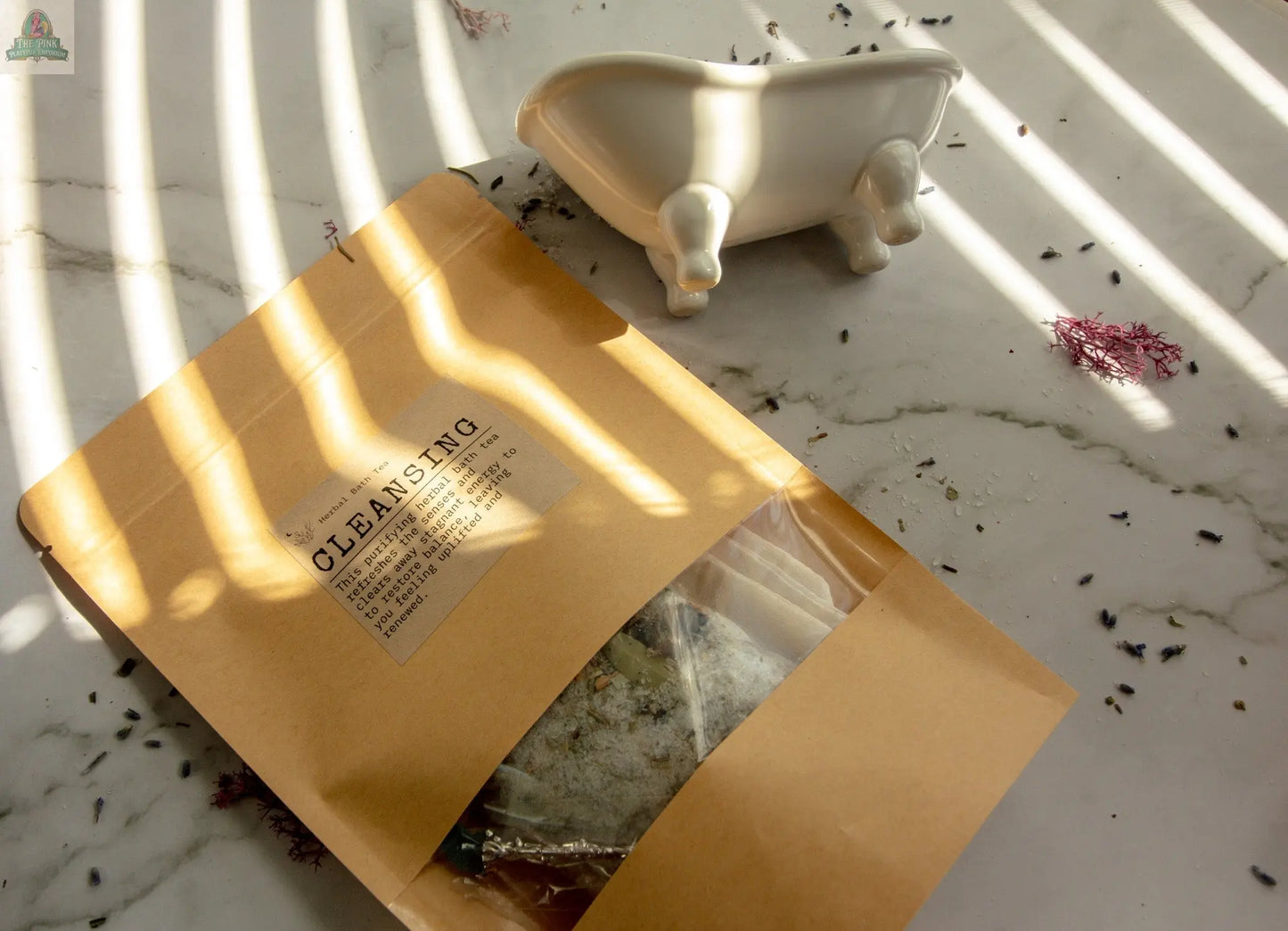 A brown paper packet labeled "Cleansing" herbal bath tea sits on a white marble surface near an empty white bathtub-shaped dish, with sunlight casting striped shadows for a serene self-care vibe.