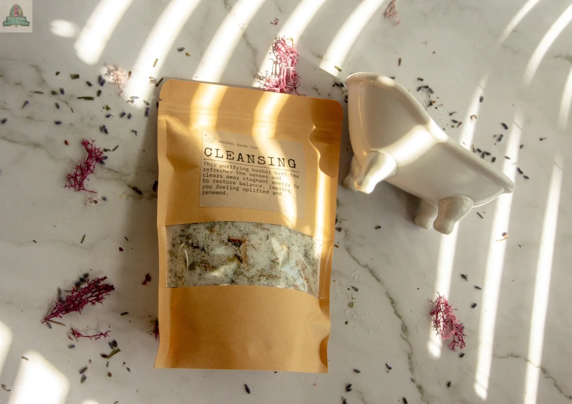 A brown resealable pouch labeled Cleansing rests on a marble surface with scattered herbs; to the right, a small ceramic bathtub—ideal for an herbal bath tea self-care ritual—sits partly in sunlight and shadow.