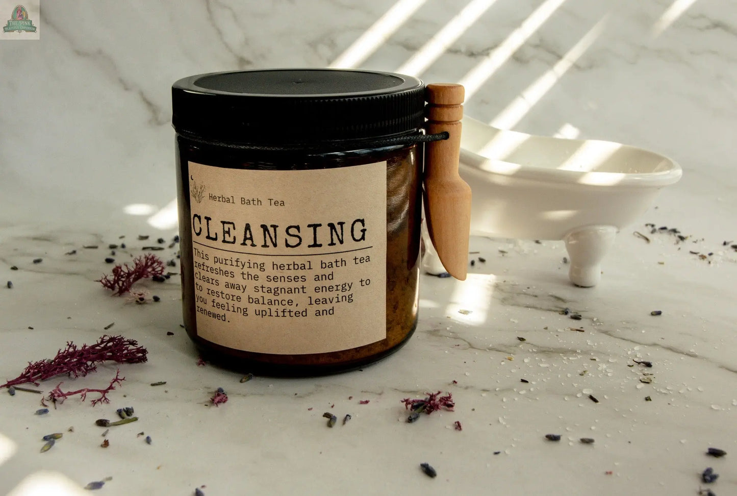A jar of Cleansing herbal bath tea rests on marble with a wooden scoop. Dried herbs, a small white dish, and subtle essential oil hints set a soothing self-care mood, sunlight filtering through the blinds.