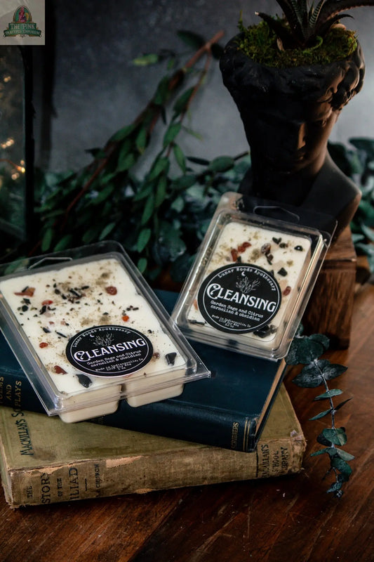 Two “Cleansing — Witchy Wax Melts” with herbs and stones sit on vintage books beside a black statue planter holding a succulent, ideal for energy cleansing, with fresh greenery in the background.