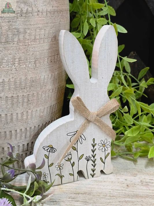 The Clover Bunny 7in figurine, featuring floral details and a twine bow, sits beside a rustic pot and green foliage.