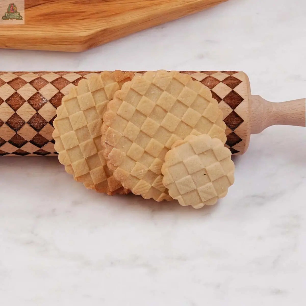 Three round cookies with a crisscross pattern rest on a decorative Courtly Check rolling pin, set on a white marble surface—the perfect way to showcase your favorite baking tools.