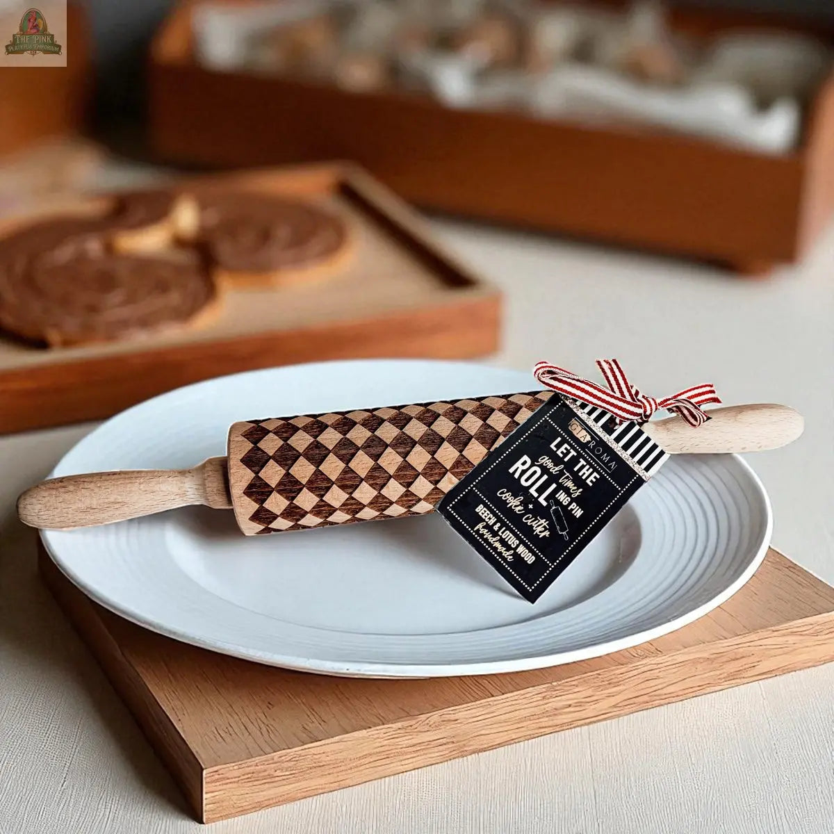 A Courtly Check patterned wooden rolling pin with a decorative tag sits on a white plate atop a wooden board, surrounded by cookies with chocolate spread and baking tools for a warm, inviting baking scene.
