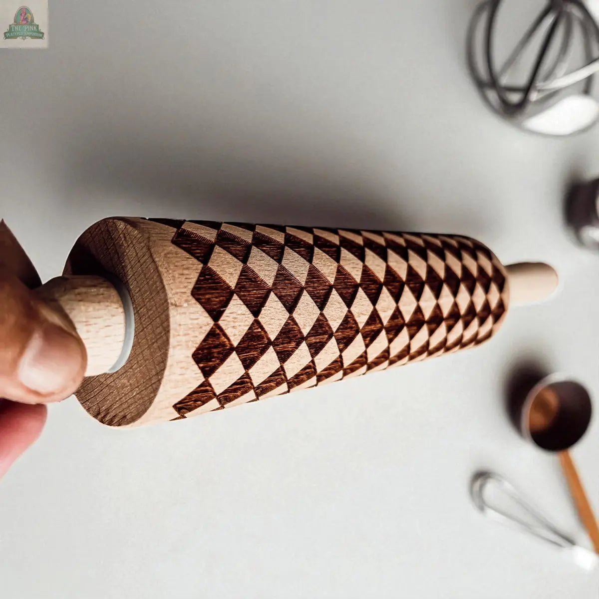 A hand holds a wooden rolling pin with a geometric diamond pattern, reminiscent of the Courtly Check collection; baking tools such as whisks, cookie cutters, and measuring cups are softly blurred in the background.