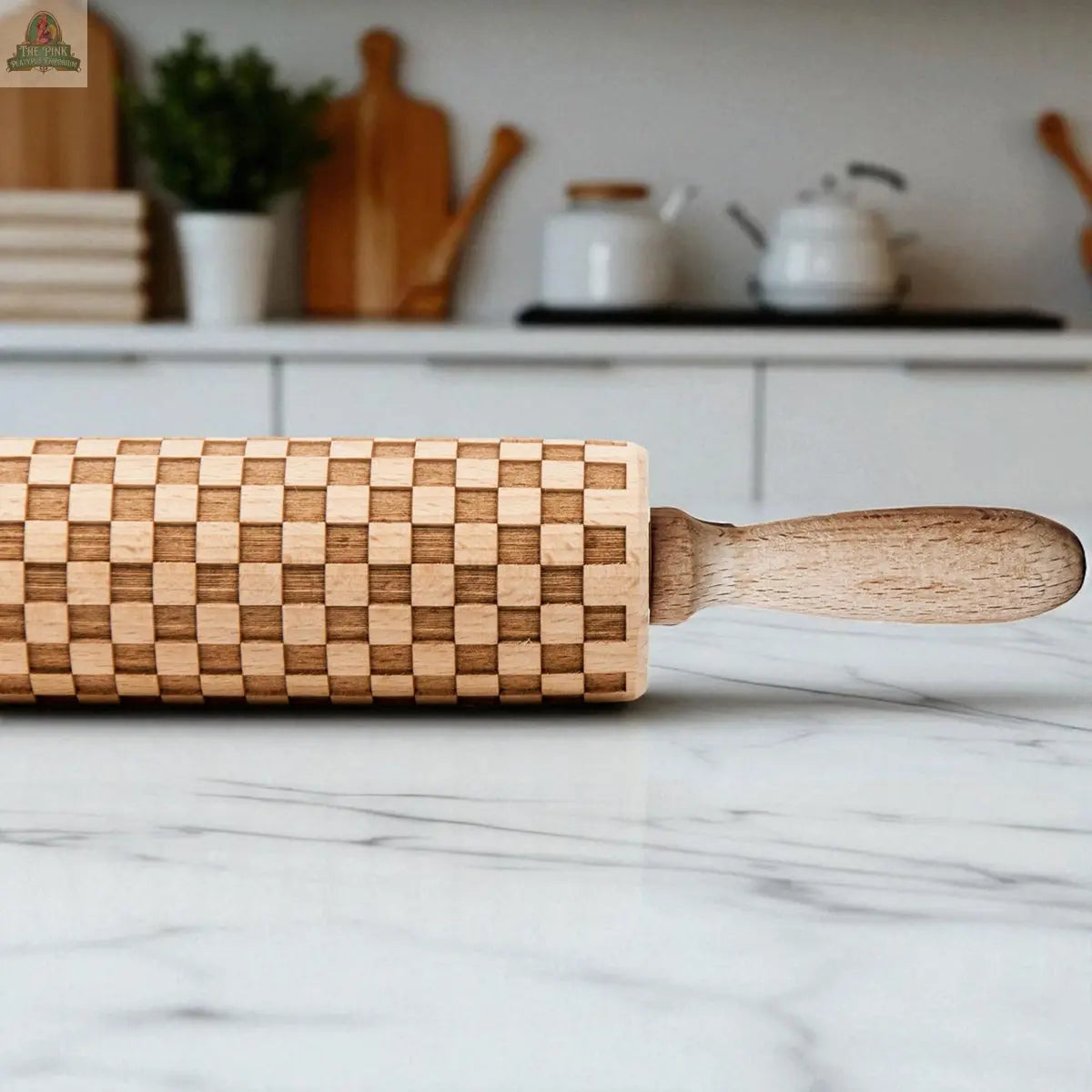 A Courtly Check rolling pin sits on a marble countertop in a modern kitchen with white cabinets, baking tools, and wooden cutting boards in the background.