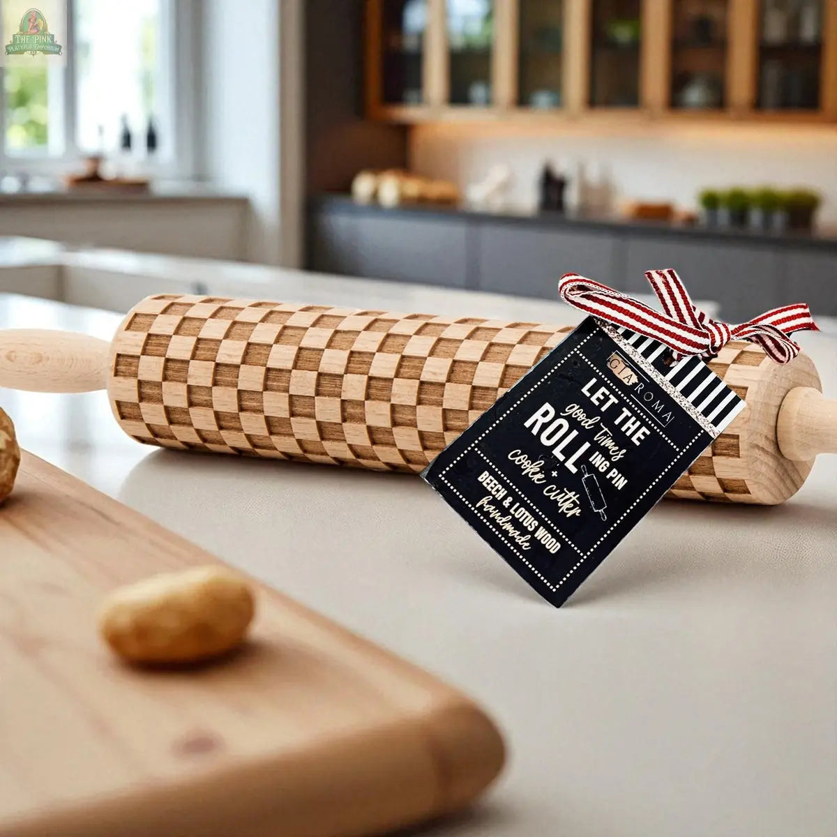 A Courtly Check rolling pin with a striped ribbon and tag rests on a kitchen countertop with baking tools and dough, set against modern cabinets and a window for a stylish, inviting atmosphere.