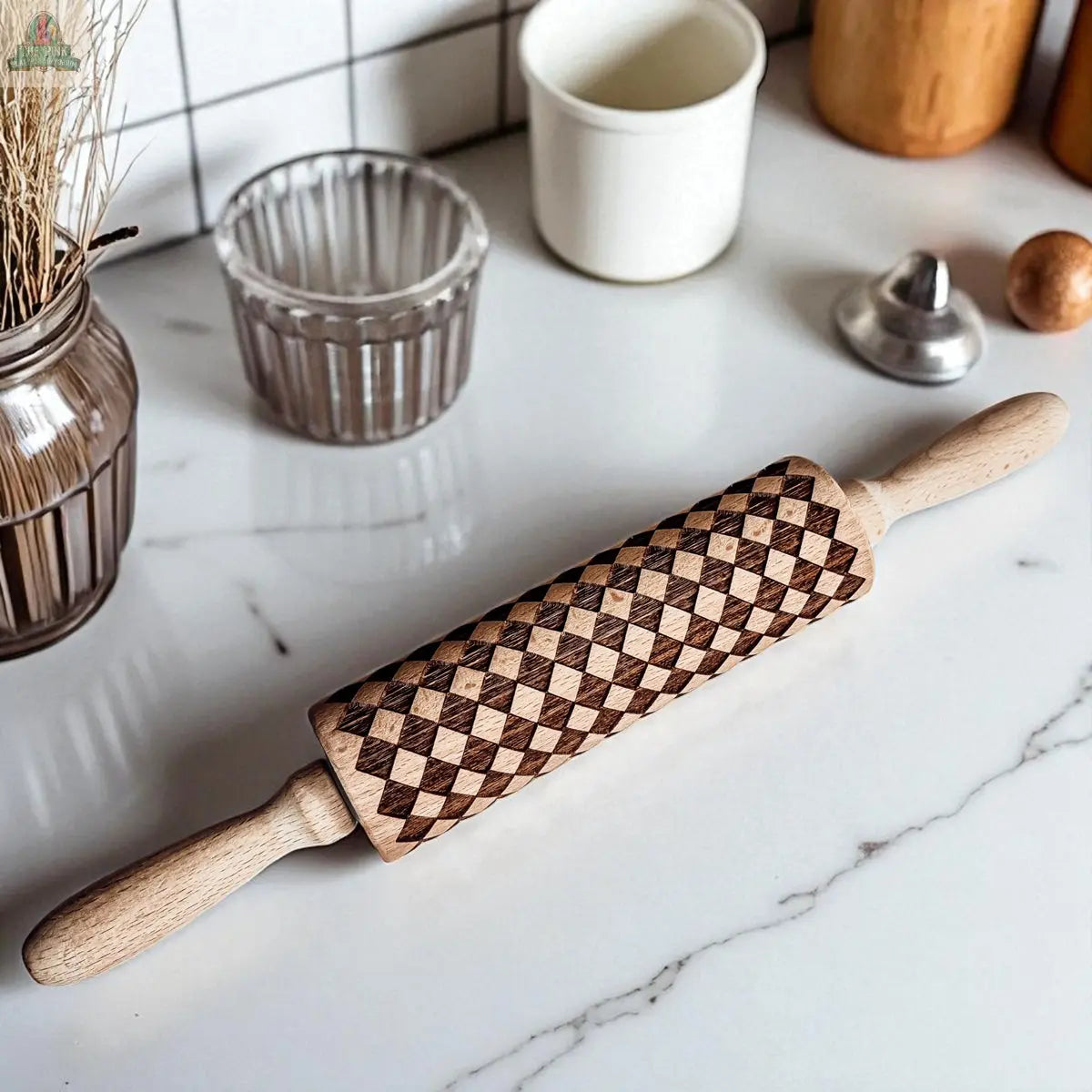 The Courtly Check rolling pin, engraved with a geometric diamond pattern, sits on a white marble countertop among baking tools like cookie cutters, jars, a glass bowl, and a small white container.