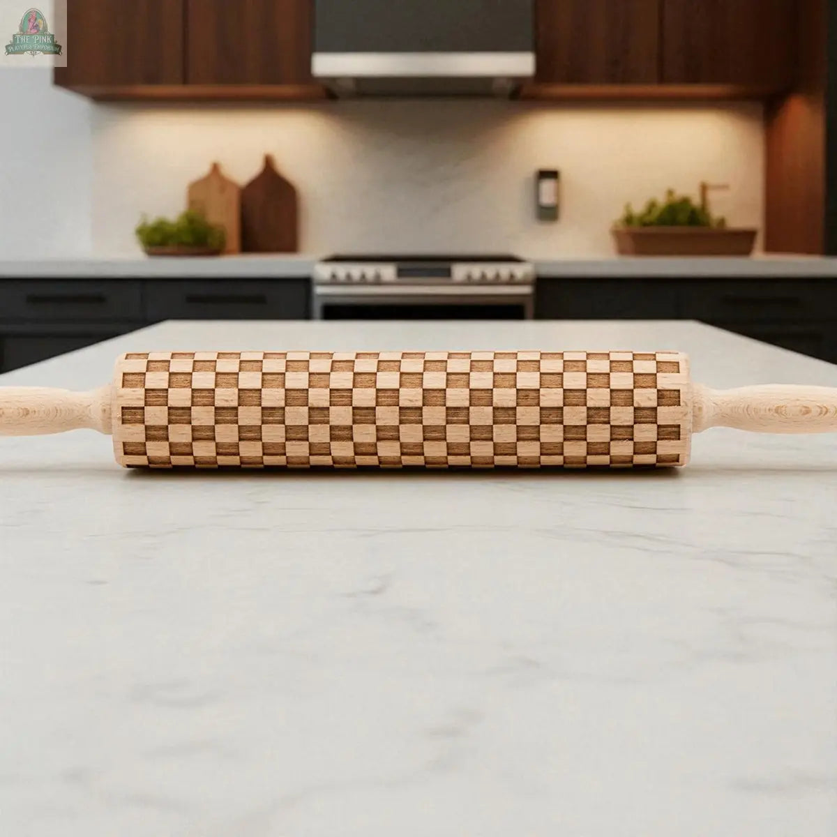 A Courtly Check rolling pin with its bold check pattern sits on a marble countertop in a modern kitchen, surrounded by dark cabinets, baking tools, plants, and cutting boards.
