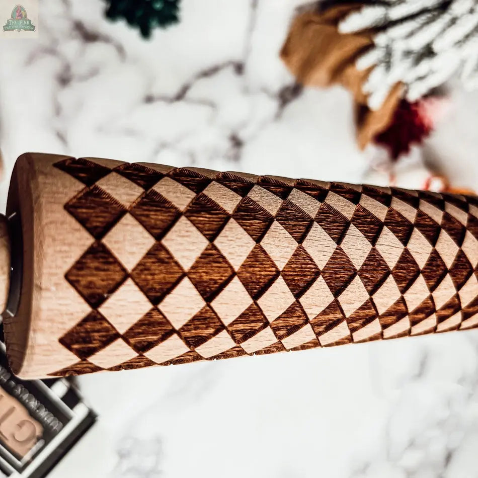 A close-up of the Courtly Check rolling pin, featuring a geometric diamond pattern, set against a blurred background with festive holiday décor and classic baking tools.