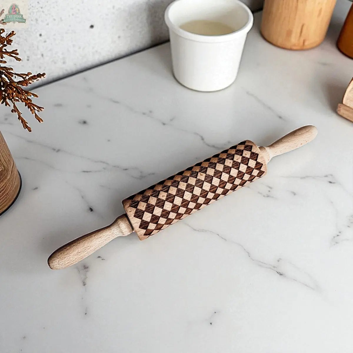 A Courtly Check rolling pin with its signature dark and light pattern sits on a white marble countertop in the kitchen, surrounded by baking tools and charming decor.