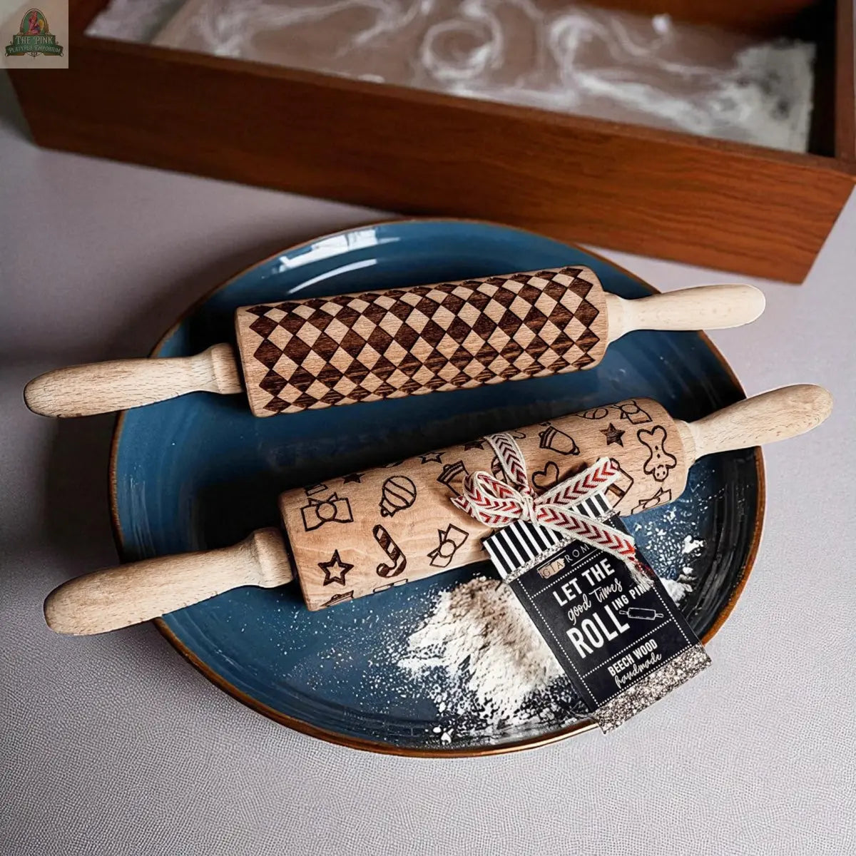 Two engraved wooden rolling pins, including the Courtly Check design, rest on a blue plate dusted with flour. One features geometric diamonds; the other has baking icons. A tag is attached—perfect for pairing with cookie cutters.