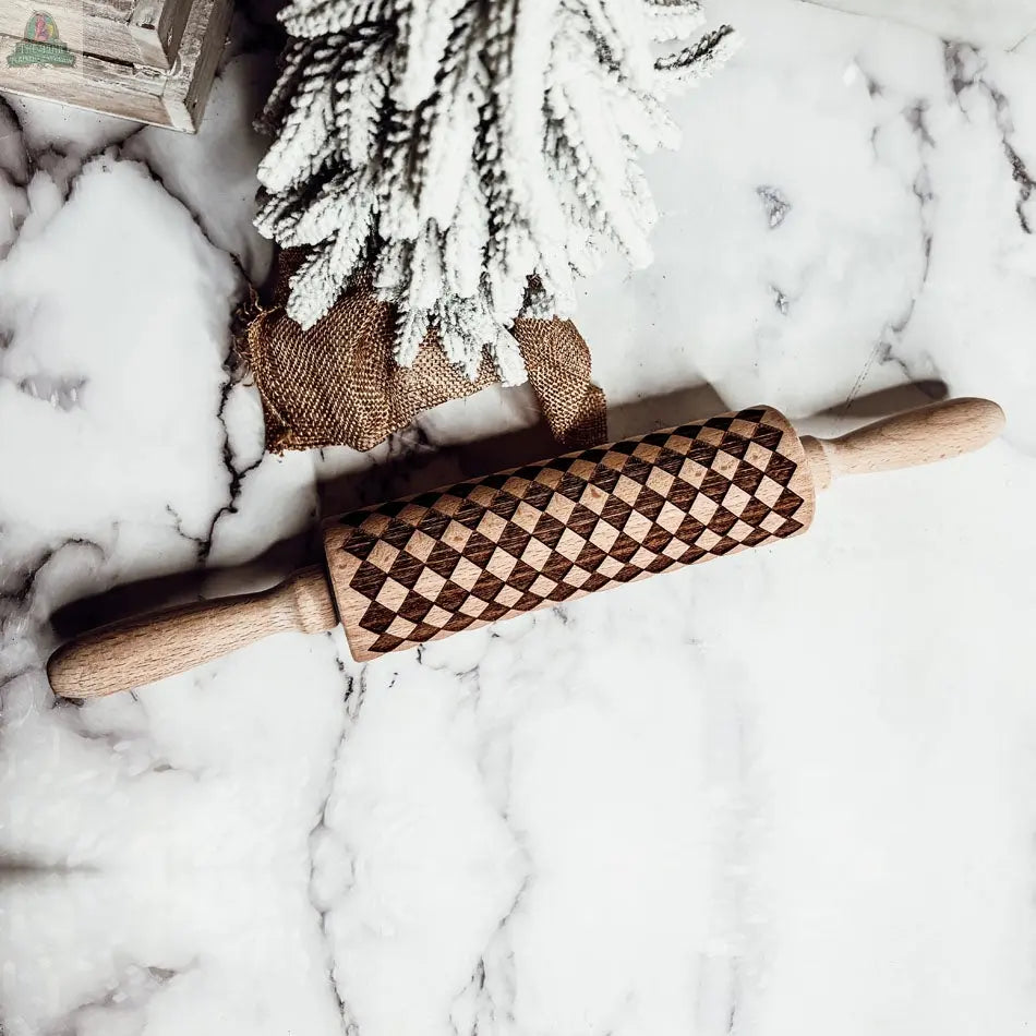 A Courtly Check rolling pin with a geometric diamond pattern rests on marble beside a snow-dusted, burlap-wrapped artificial tree—perfect for charming your baking collection.