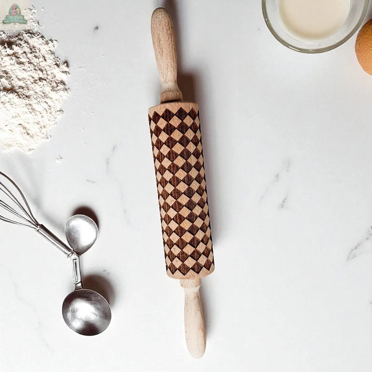 A Courtly Check rolling pin sits on a white marble surface, surrounded by a bowl of milk, flour, metal measuring spoons, and part of an egg—essential baking tools for every kitchen.