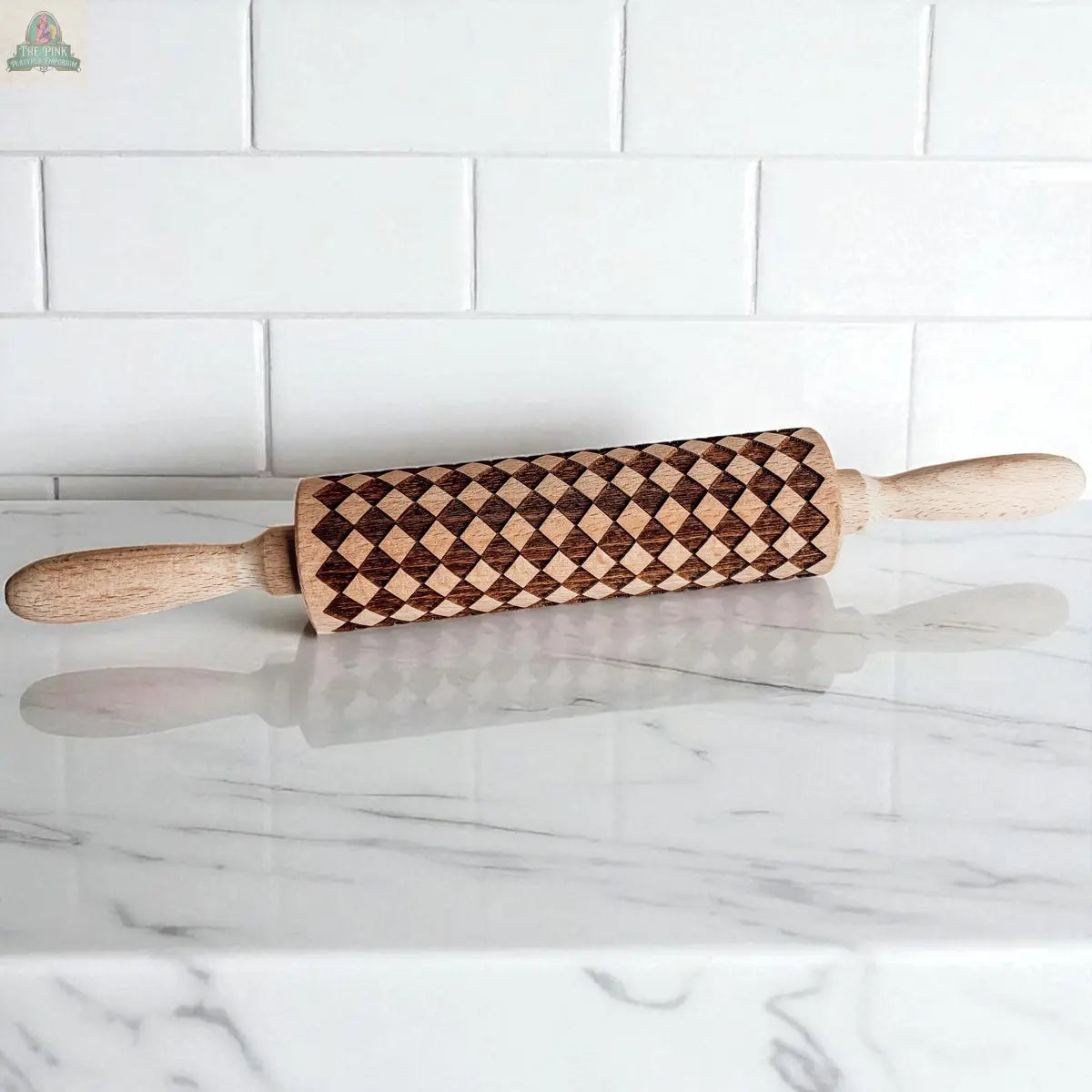 A Courtly Check rolling pin with wooden handles and a patterned cylinder featuring a dark diamond design sits among baking tools on a white marble countertop in front of a white tiled backsplash.