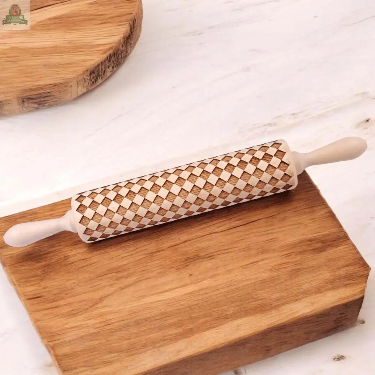 The Courtly Check wooden rolling pin, featuring a diamond-carved pattern, sits on a wooden board with a round board nearby, all arranged on a light marble surface—ideal for upgrading your baking tools collection.