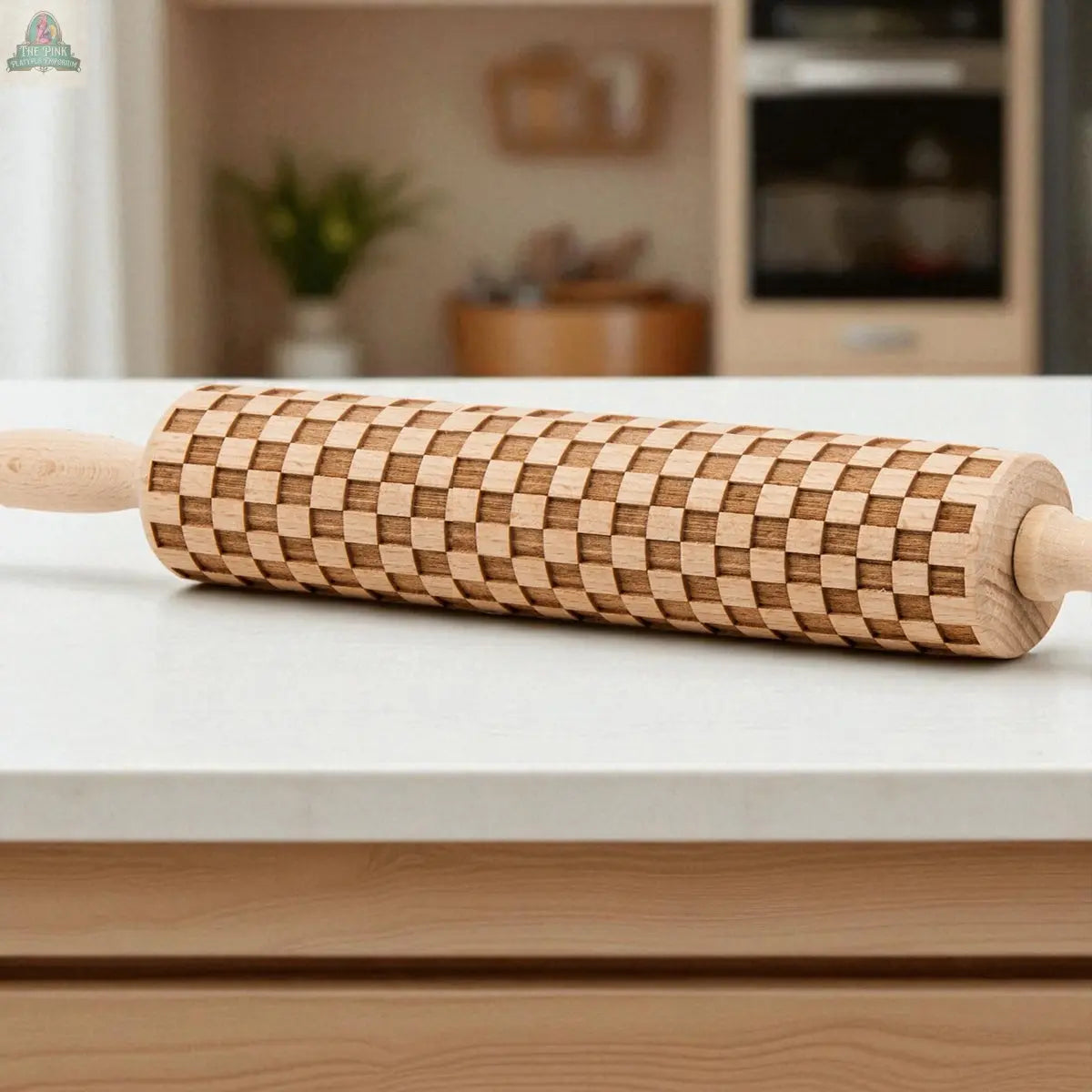 A Courtly Check rolling pin sits on a white kitchen countertop with baking tools nearby, while blurred shelves, a plant, and an oven appear in the background.