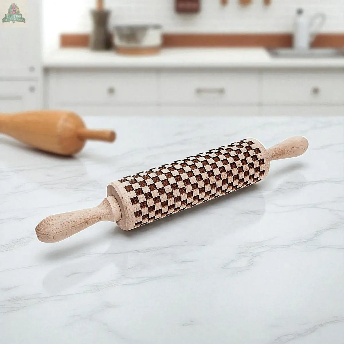 A Courtly Check rolling pin featuring a bold wood design rests on a white marble countertop, with baking tools, cookie cutters, and another blurred rolling pin in the background.