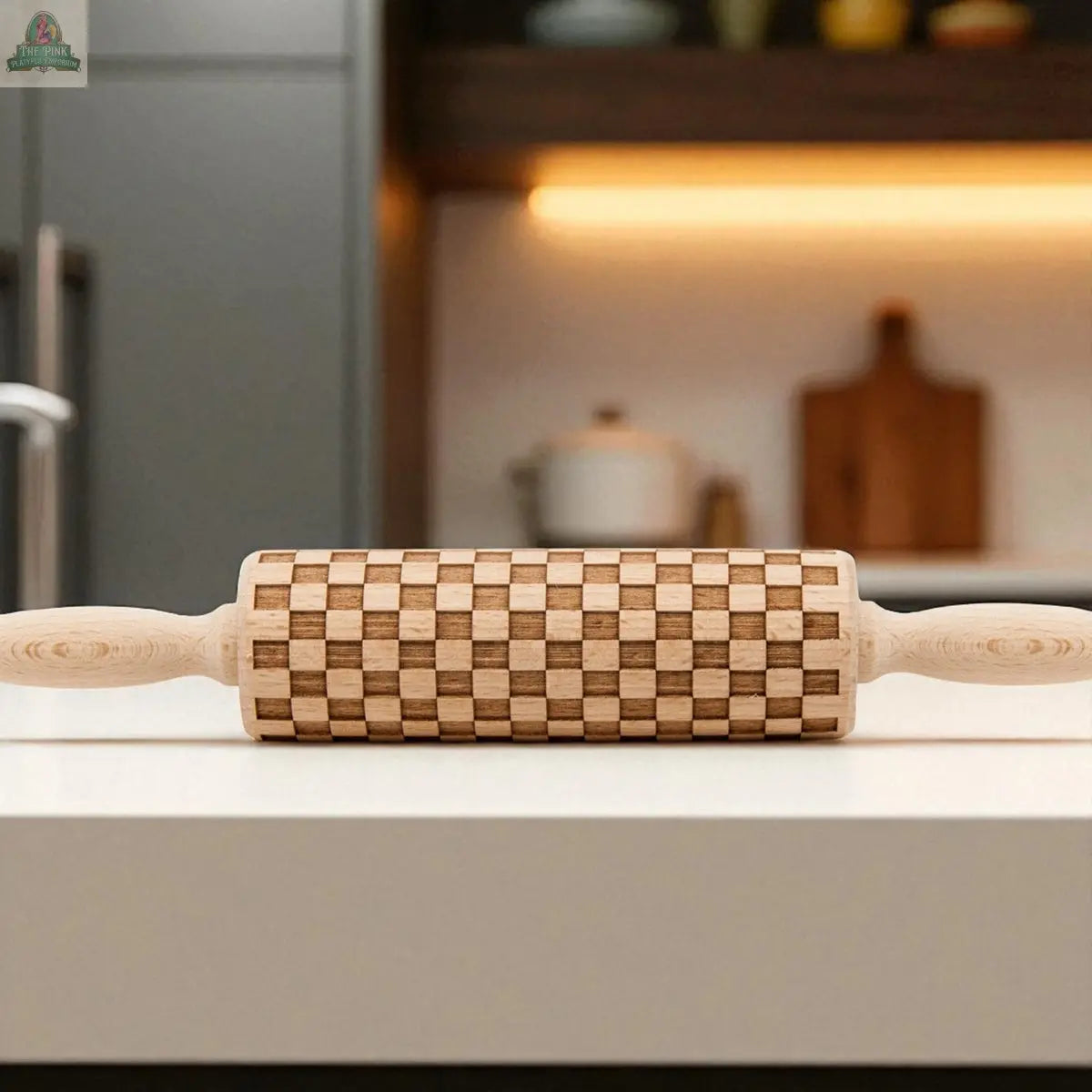A Courtly Check rolling pin with a distinctive checkerboard pattern sits on a white kitchen counter, surrounded by modern baking tools and utensils in the blurred background.