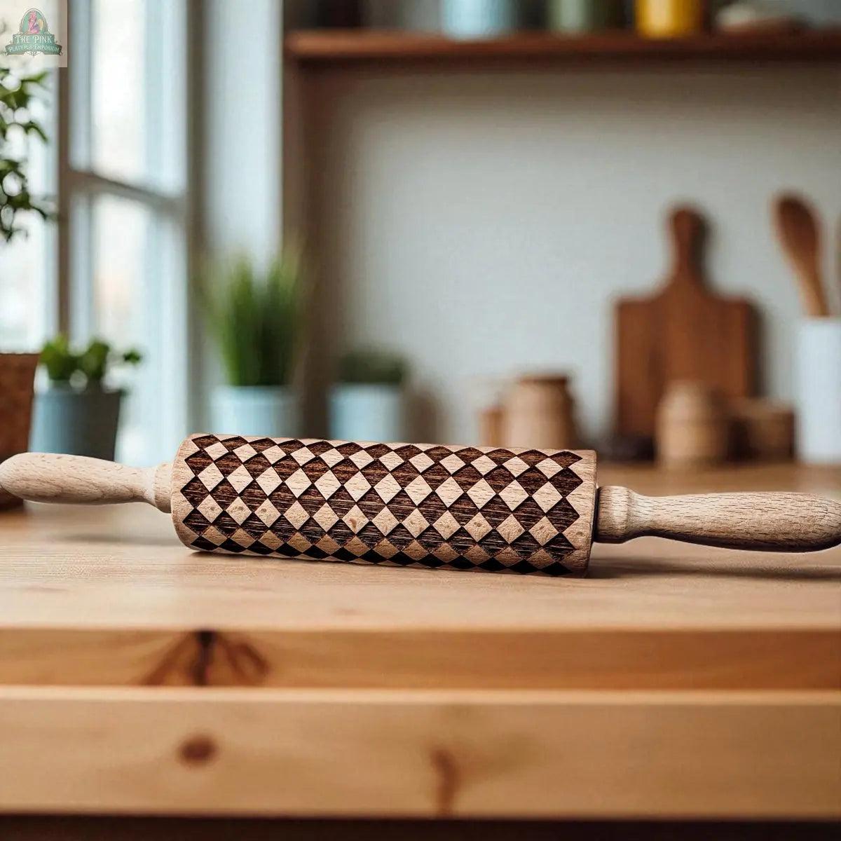 A Courtly Check wooden rolling pin with a diamond-patterned design sits on a kitchen counter, surrounded by blurred baking tools and potted plants, adding charm to your kitchen décor.