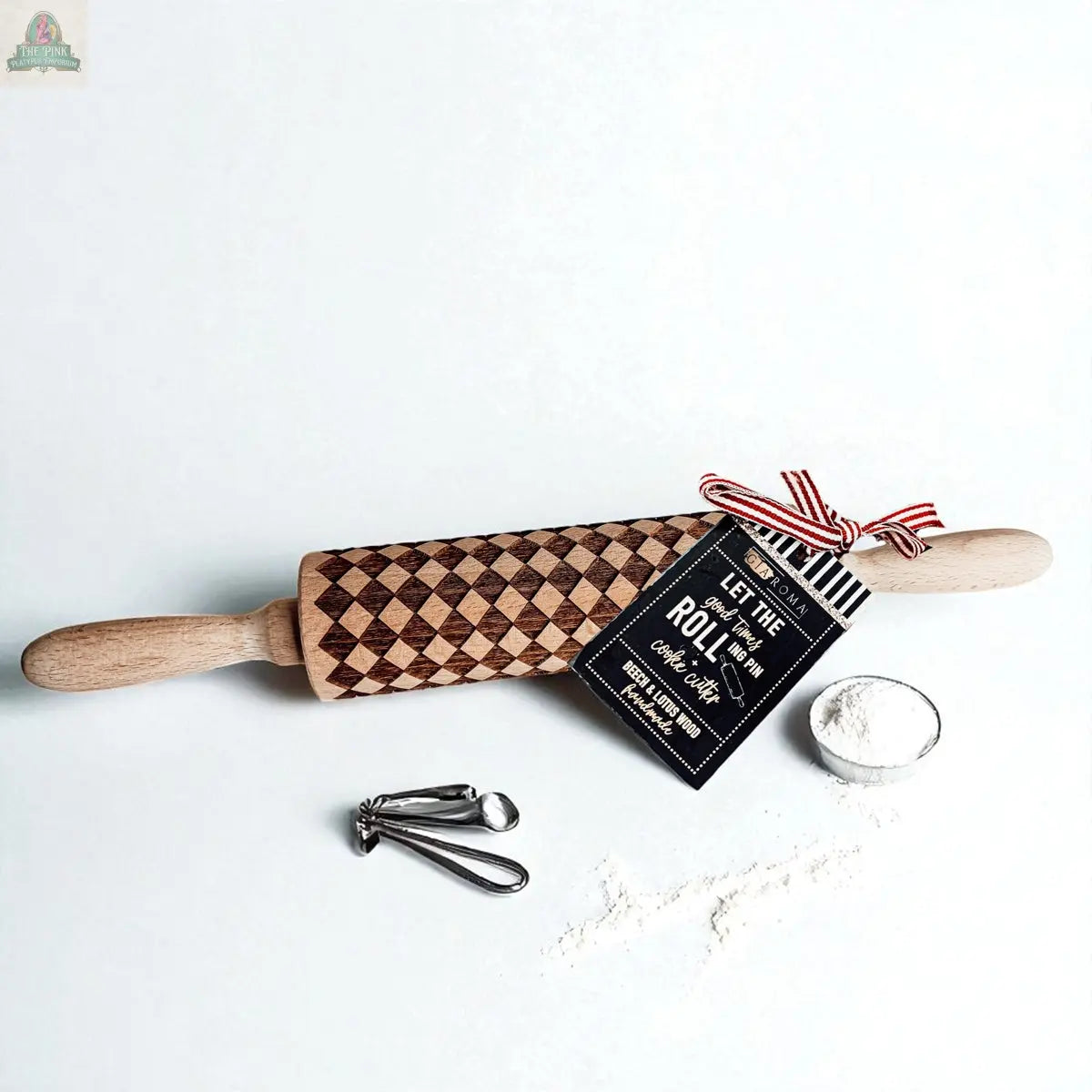 A Courtly Check rolling pin rests on a white surface beside flour, metal measuring spoons, and a recipe card tied with a red-striped ribbon.