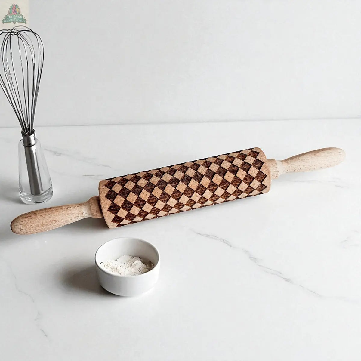 The Courtly Check patterned wooden rolling pin with light handles sits on a white countertop beside a small bowl of flour, a metal whisk in a stand, and assorted cookie cutters—an inviting display of classic baking tools.