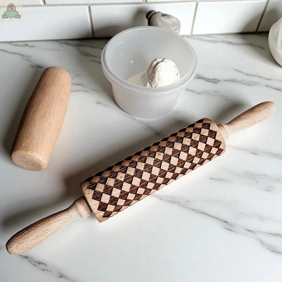 The Courtly Check patterned wooden rolling pin with its signature geometric design rests on a marble countertop beside a plain wooden dough roller, essential baking tools, and a plastic container holding dough, all set against a white tile backsplash.