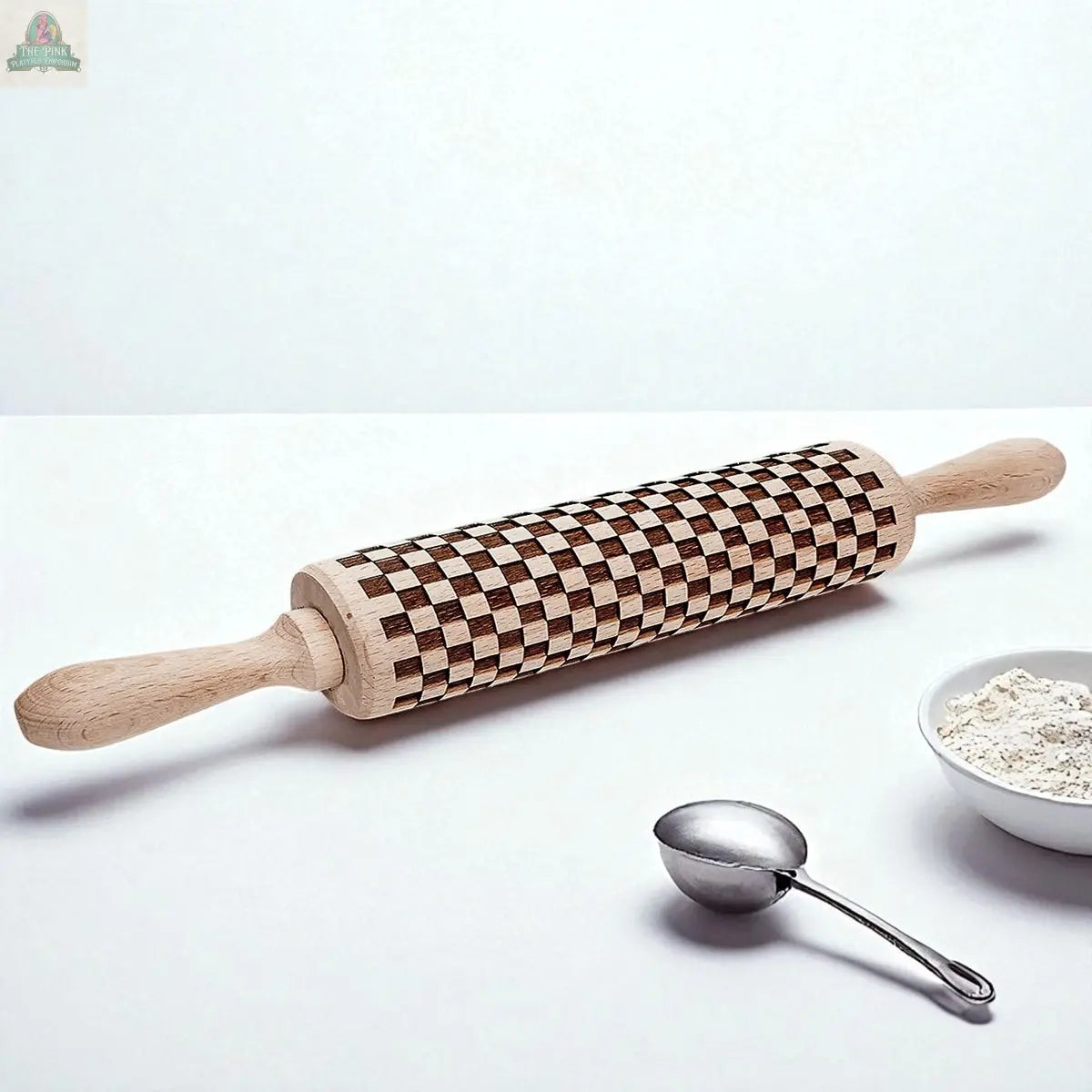 A Courtly Check rolling pin with a brown and beige checkerboard pattern rests on a white surface beside a metal measuring spoon and a bowl of flour—perfect for your baking tools collection.