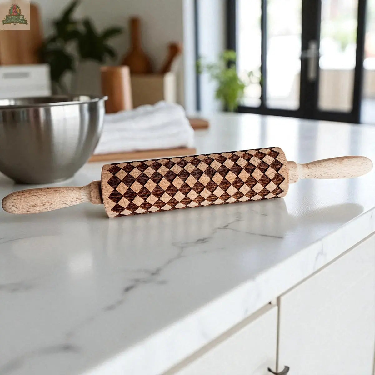 A Courtly Check rolling pin featuring a geometric diamond pattern sits on a white marble countertop, surrounded by a metal mixing bowl, cookie cutters, and various baking tools.
