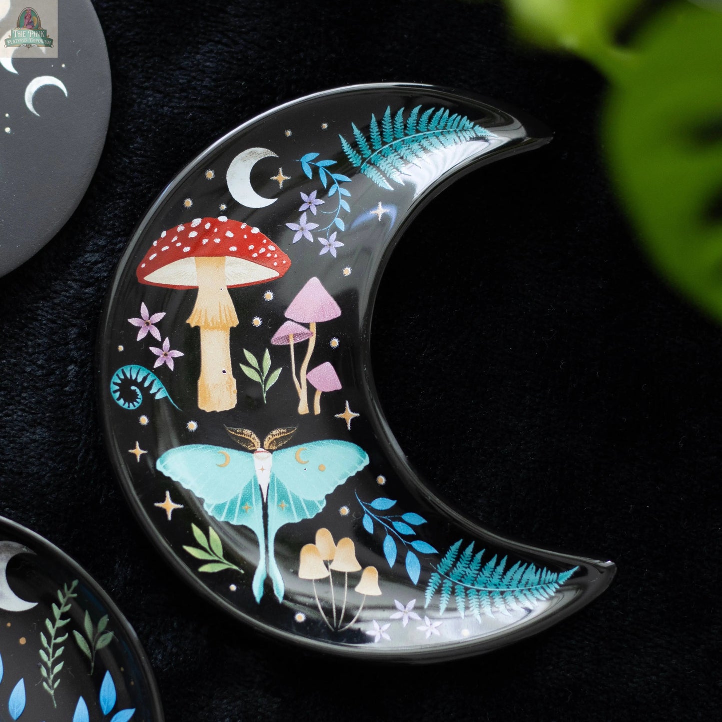 The Crescent Moon Dark Forest Print Trinket Dish features pastel mushrooms, ferns, stars, and a moth on a black base. This celestial decor piece serves as a jewellery holder and is pictured resting on a dark surface with a green leaf nearby.