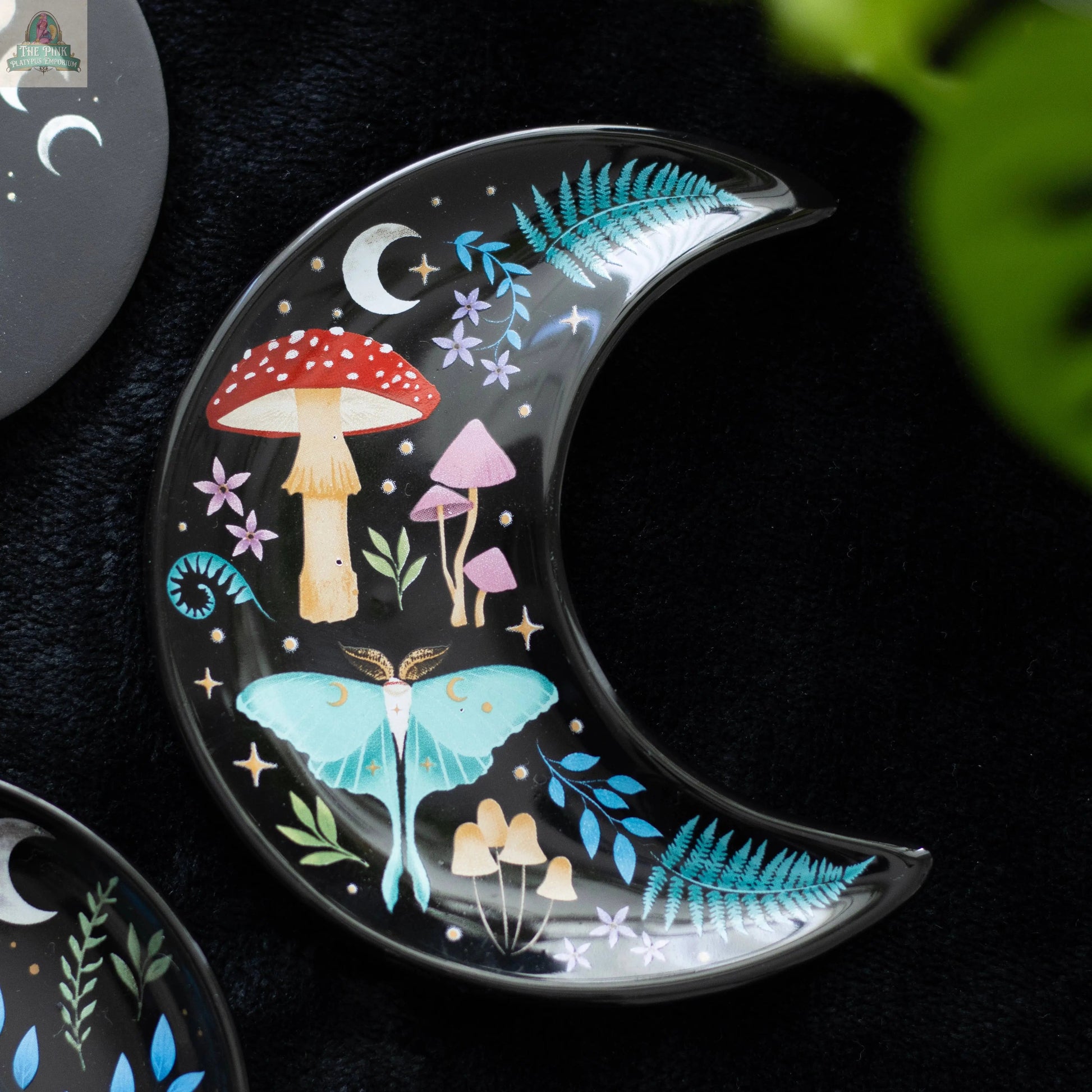 The Crescent Moon Dark Forest Print Trinket Dish features pastel mushrooms, ferns, stars, and a moth on a black base. This celestial decor piece serves as a jewellery holder and is pictured resting on a dark surface with a green leaf nearby.