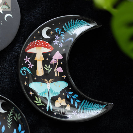 The Crescent Moon Dark Forest Print Trinket Dish features pastel mushrooms, ferns, stars, and a moth on a black base. This celestial decor piece serves as a jewellery holder and is pictured resting on a dark surface with a green leaf nearby.