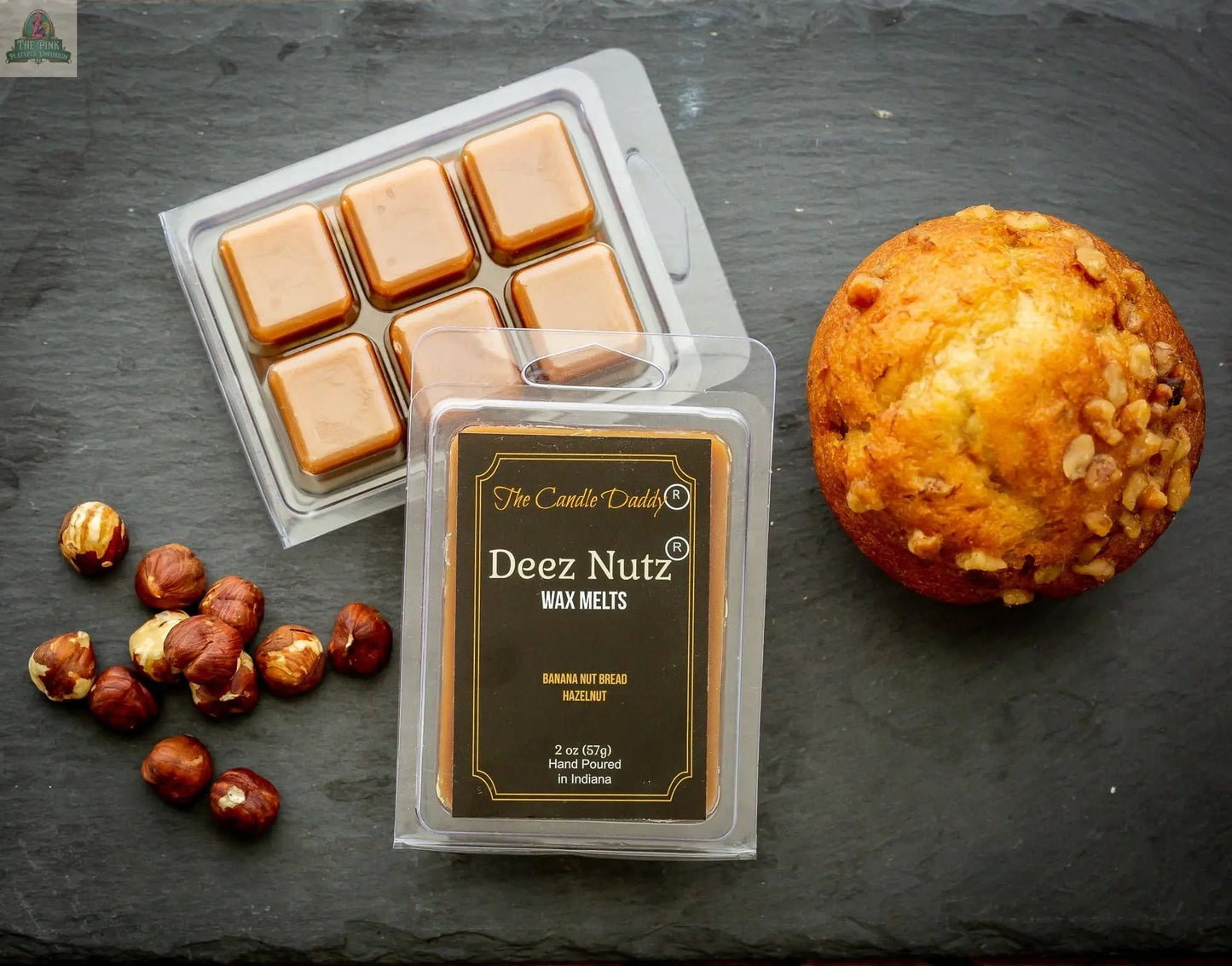 A muffin, hazelnuts, caramel squares, and a pack of Deez Nutz Wax Melts - Banana Nut Bread are displayed on a dark slate surface.