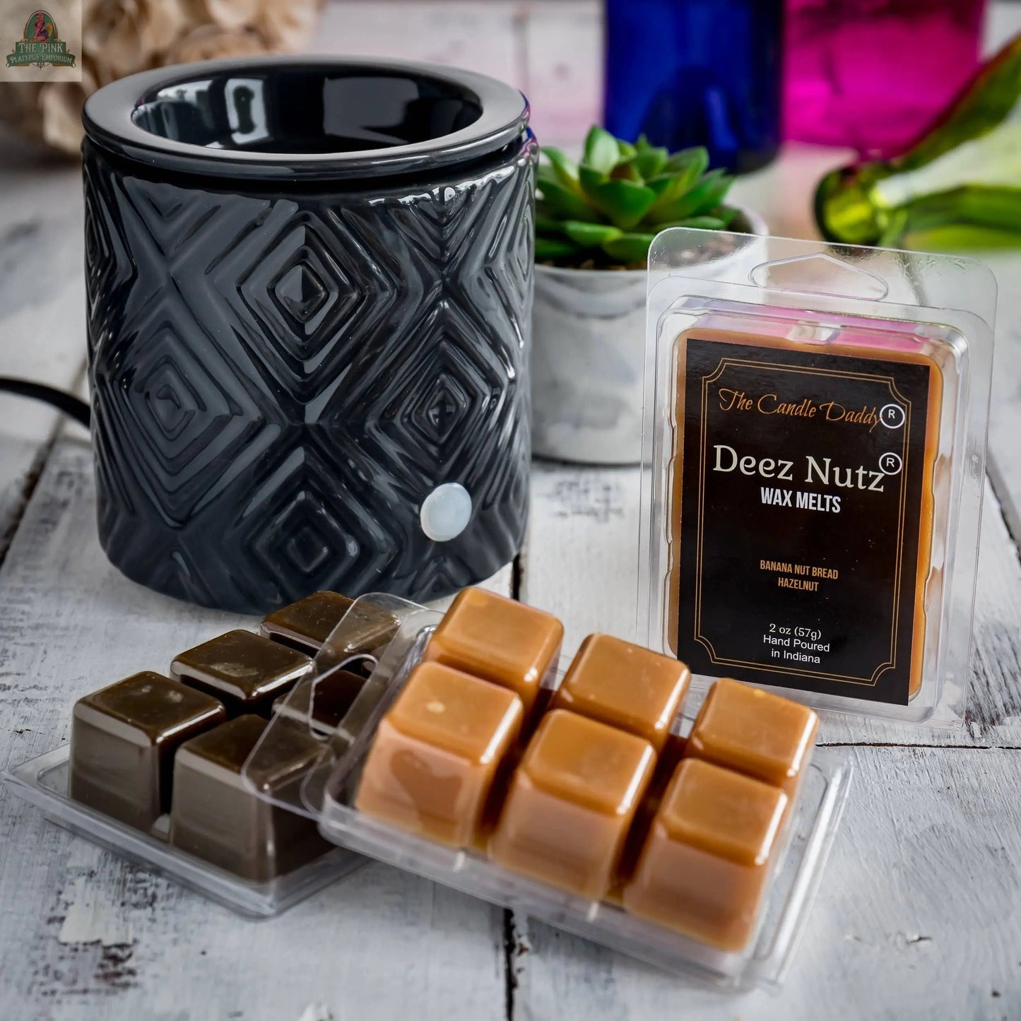 A black geometric wax warmer sits on a white wooden surface next to an open pack of Deez Nutz Wax Melts - Banana Nut Bread: 1 Pack, with plants and colorful bottles in the background.