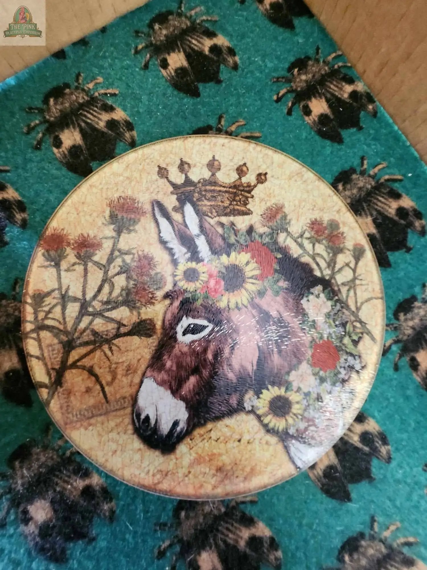 The "Donkey Crown" 12" round glass cutting board features a handmade Texas design with a donkey in a floral crown, a golden crown above, thistle plants, and is displayed on teal bee-patterned fabric—a unique accent for any charcuterie spread.