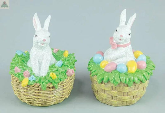 The Easter Bunny in Woven Basket 2 Ast features two white ceramic bunnies—one standing, one sitting with a pink ribbon—in cube baskets (3.54x3.54x4.53 in) filled with grass and eggs, all set against a light gray background.