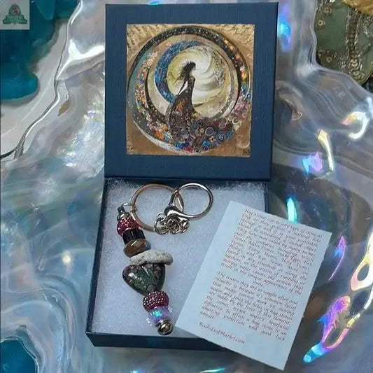 The Elegant Multicolor Beaded Hagstone Keychain comes in a blue Goddess Box with an artistic lid depicting a vibrant woman. A note with red text is included beside the keychain inside the box.