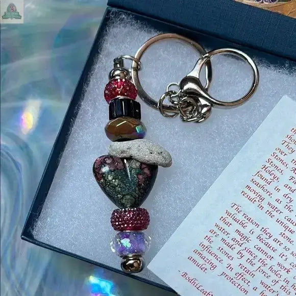 The Elegant Multicolor Beaded Hagstone Keychain with Goddess Box features a heart-shaped hagstone, colorful beads, and a silver ring, beautifully presented in a blue box with textured white lining and a partially visible note.